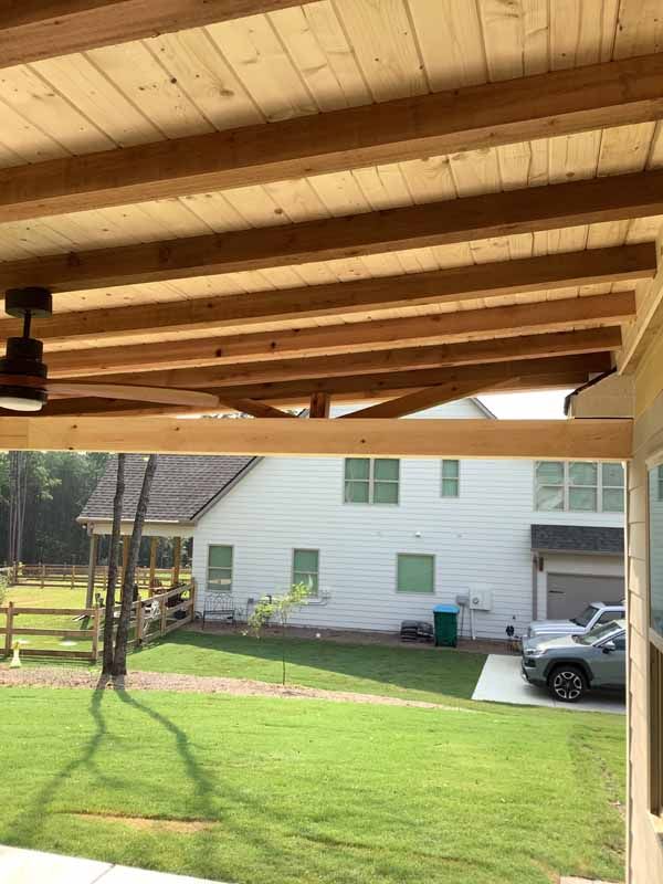 Wooden patio roof with fan, overlooking a yard and two-story house.