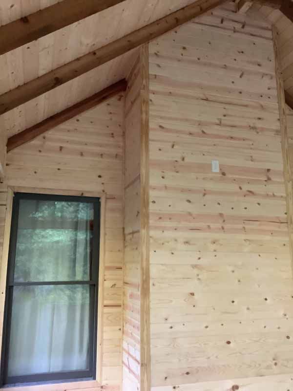 Interior of a cabin with unfinished wood paneling, a window, and exposed wooden beams.