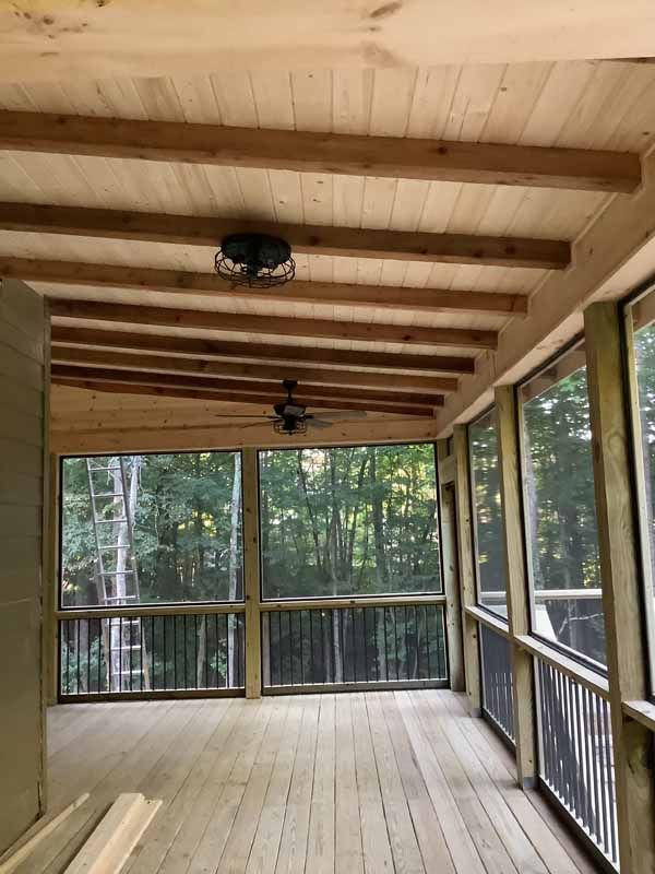 Screened-in porch with wood ceiling and floor, overlooking a forested view. A ceiling fan hangs in the center.
