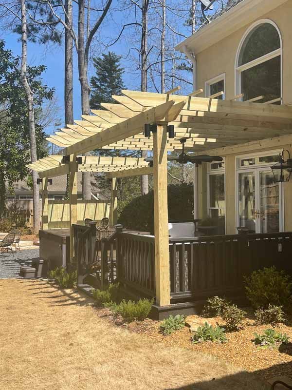 Wooden pergola on a deck attached to a light-colored house. Sunny day with trees in the background.