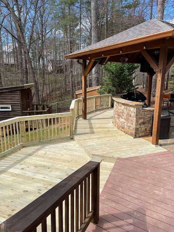 Wooden deck with a gazebo, built-in grill, and a brown railing, set against a wooded backdrop.