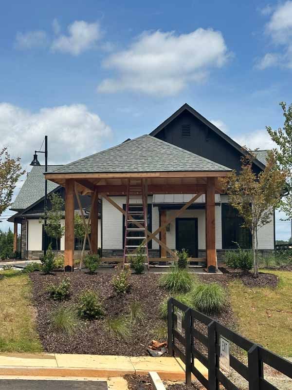 Wooden structure with a gray shingled roof, built in front of a modern white building with black accents, blue sky.