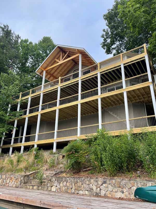 Three-story house with multiple wooden decks, surrounded by trees. Stone retaining wall in the foreground.