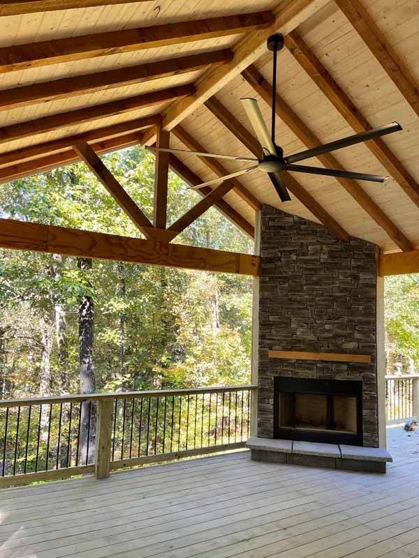 Covered deck with fireplace, exposed wooden beams, large ceiling fan, and forest view.