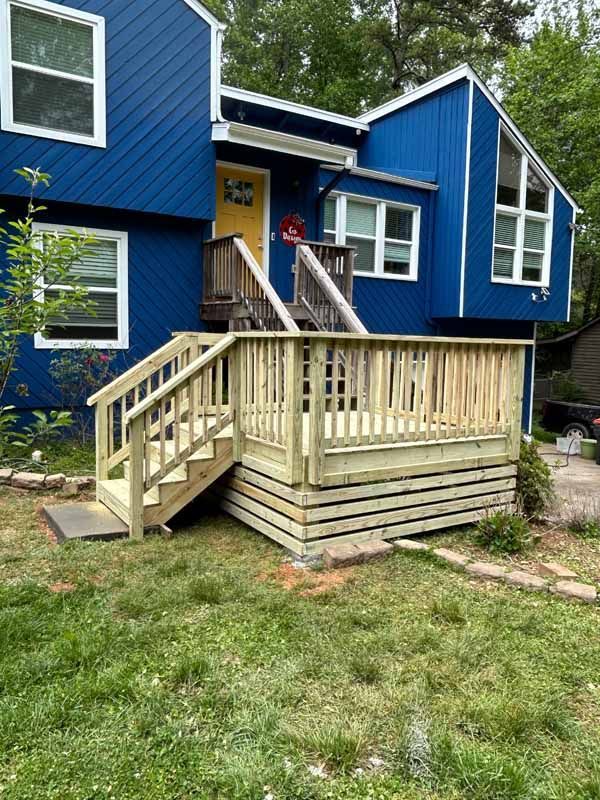 A two-story blue house with a yellow front door, featuring a new, light-wood deck with stairs in the front yard.