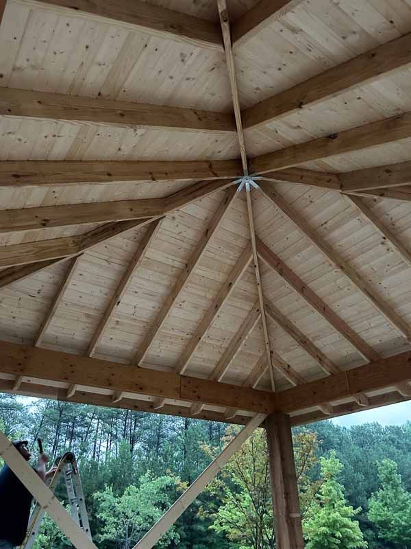 Underneath a wooden pavilion roof, a person on a ladder works on the structure's framing.