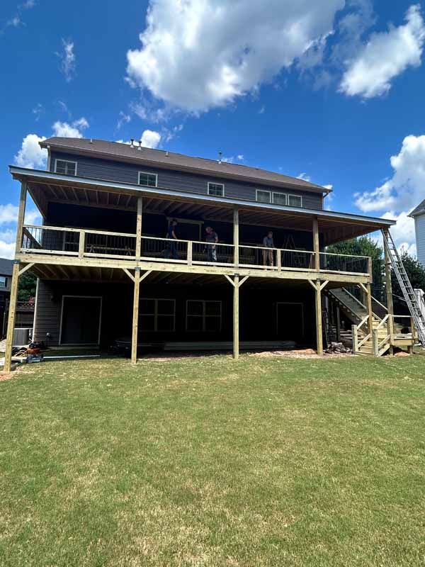 Two-story house with large wooden deck; brown siding and roof; blue sky with clouds; green grass in yard.
