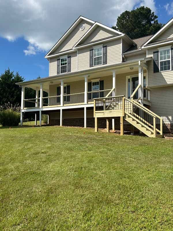 Two-story beige house with a wraparound porch, wooden deck, and green lawn under a blue sky.