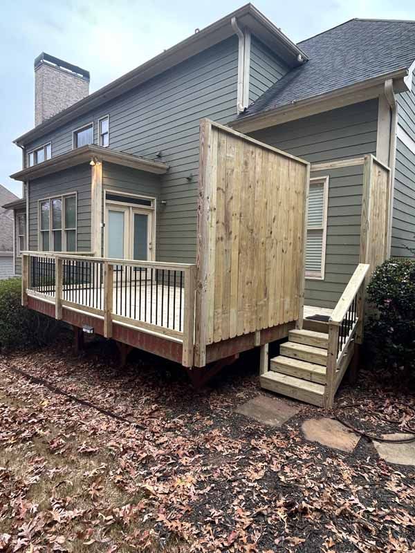 A wooden deck with metal railings and a privacy wall attached to the side of a grey two-story house with a chimney.