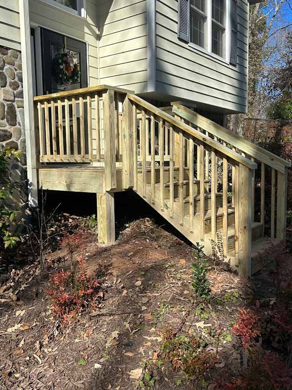 Wooden stairs with railing leading to a house entrance. Surrounding landscaping of shrubs and mulch.