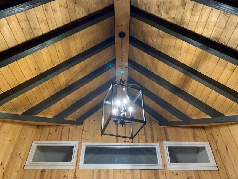 Wooden cabin interior with windows, sloped wood ceiling, black beams, and a lantern-style light fixture.