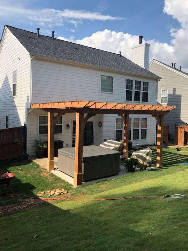 Backyard with a hot tub under a wooden pergola, next to a two-story white house with a green lawn.