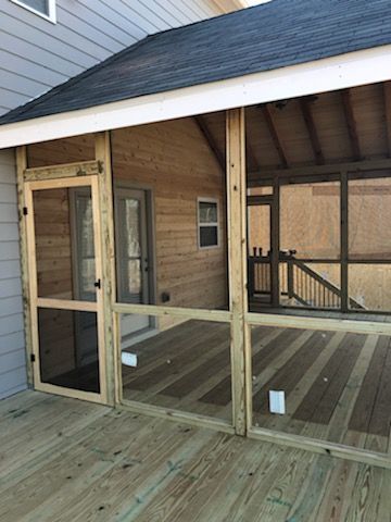 Wooden screened-in porch with door and deck. Exterior view, light wood, gray siding, black roof.