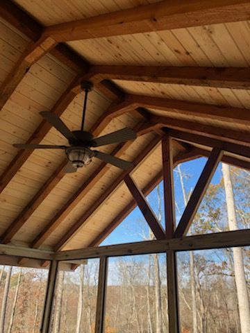 Wooden ceiling of a screened porch with a ceiling fan, triangular beams, and a view of trees and sky.