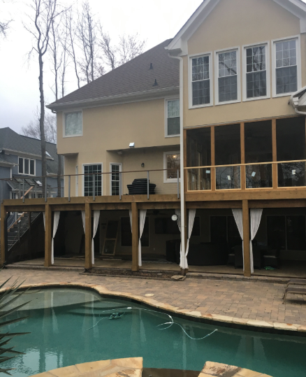 Back of a house with deck overlooking a swimming pool. Outdoor seating and curtains are beneath the deck.