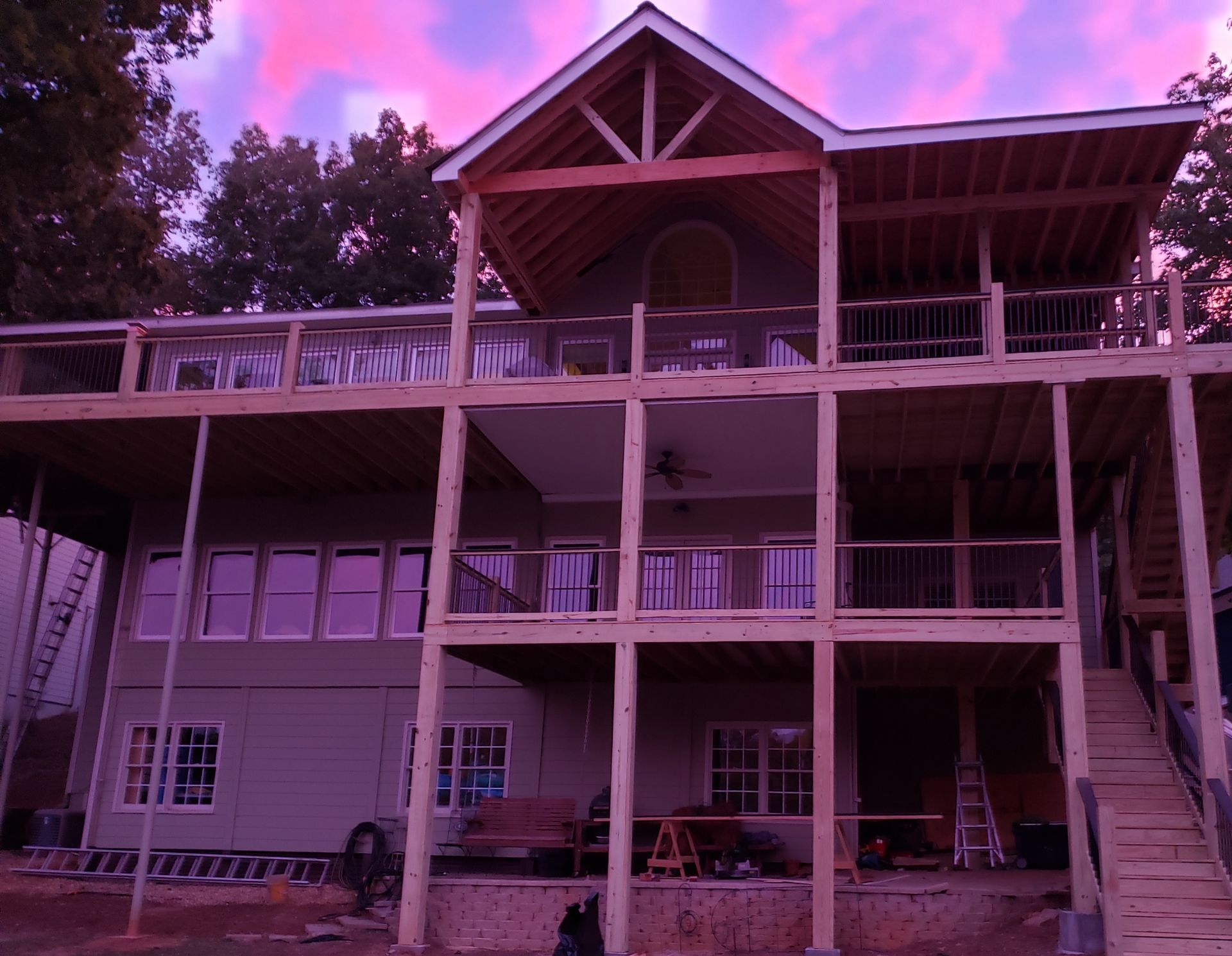 A three-story house under construction with a large wooden deck frame, green siding, and a sunset sky in the background.