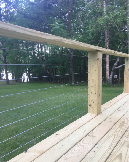 Wooden deck railing with horizontal steel cables overlooking a green lawn and trees.