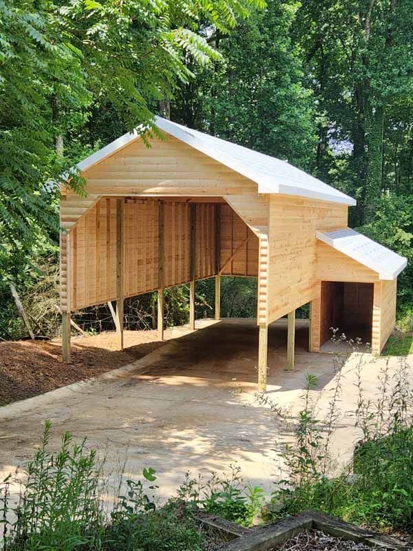 A newly constructed light-colored wooden carport with an attached small shed, built on a concrete pad in a wooded area.