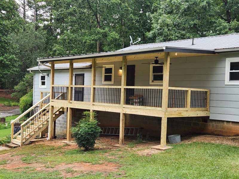 Wooden deck with railing and stairs attached to a light gray house under a metal roof, surrounded by trees.