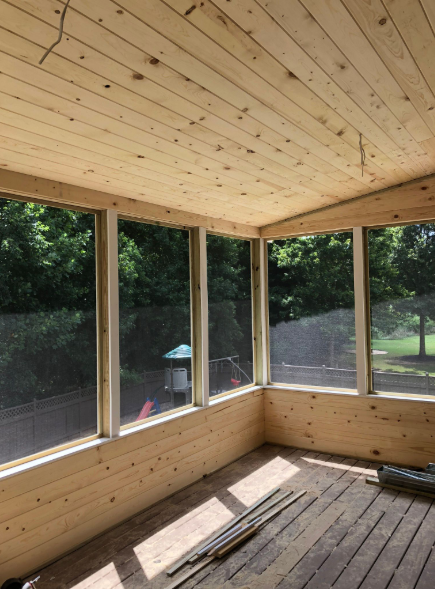 Screened-in porch with wood ceiling, walls, and screens, looking out to greenery.