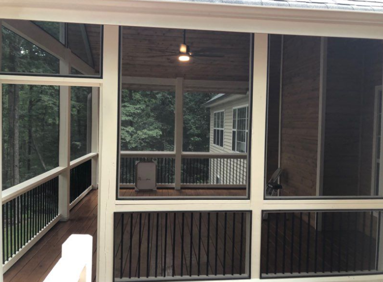Screened-in porch with dark screens, wooden railing, and a ceiling fan; view to outside greenery and a house.