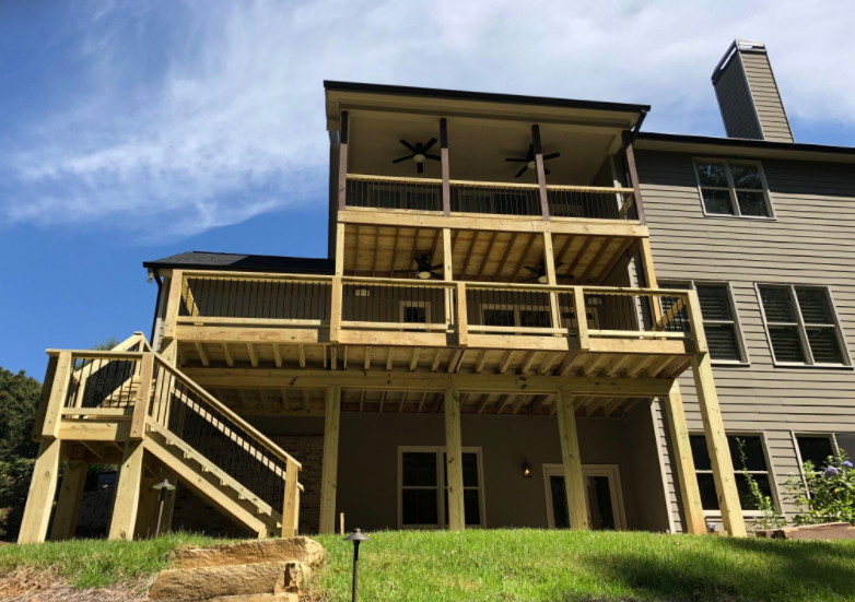 Two-story wooden deck attached to a light brown house with stairs, against a blue sky.