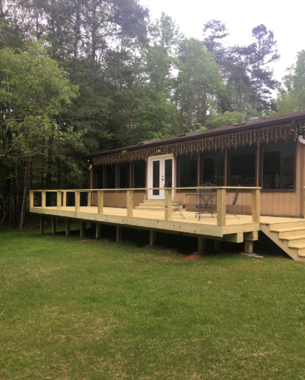 Wooden deck with railings in front of a house, set in a grassy yard, surrounded by trees.
