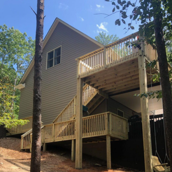 House with large wooden deck, stairs, and railing in a wooded setting under a blue sky.