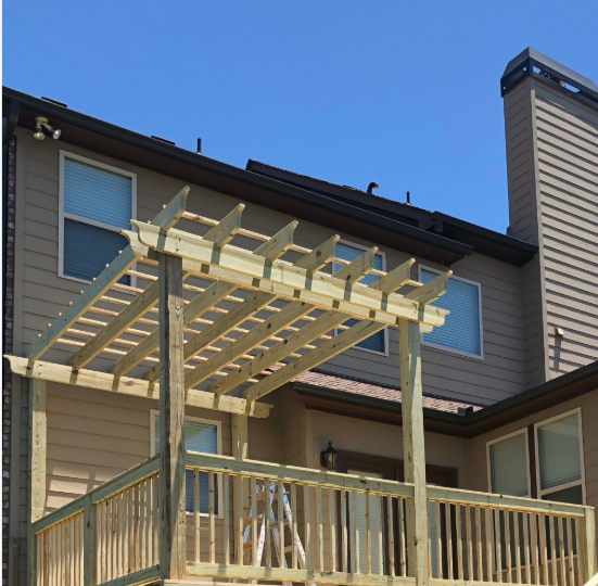 Wooden pergola built over a deck attached to a two-story beige house with blue sky.