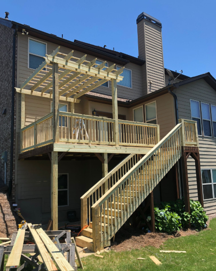Two-story wooden deck with stairs and pergola attached to a light brown house with a chimney, built in a grassy yard.