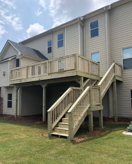 Two-story wooden deck attached to a beige house, with stairs leading down to a grassy yard under a cloudy sky.