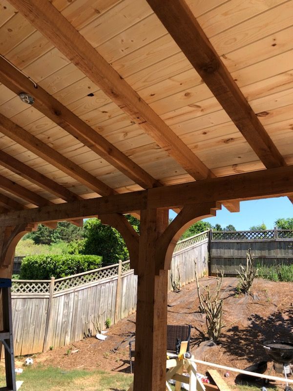 Wooden patio roof framing with exposed rafters and tongue-and-groove ceiling boards overlooking a fenced backyard.