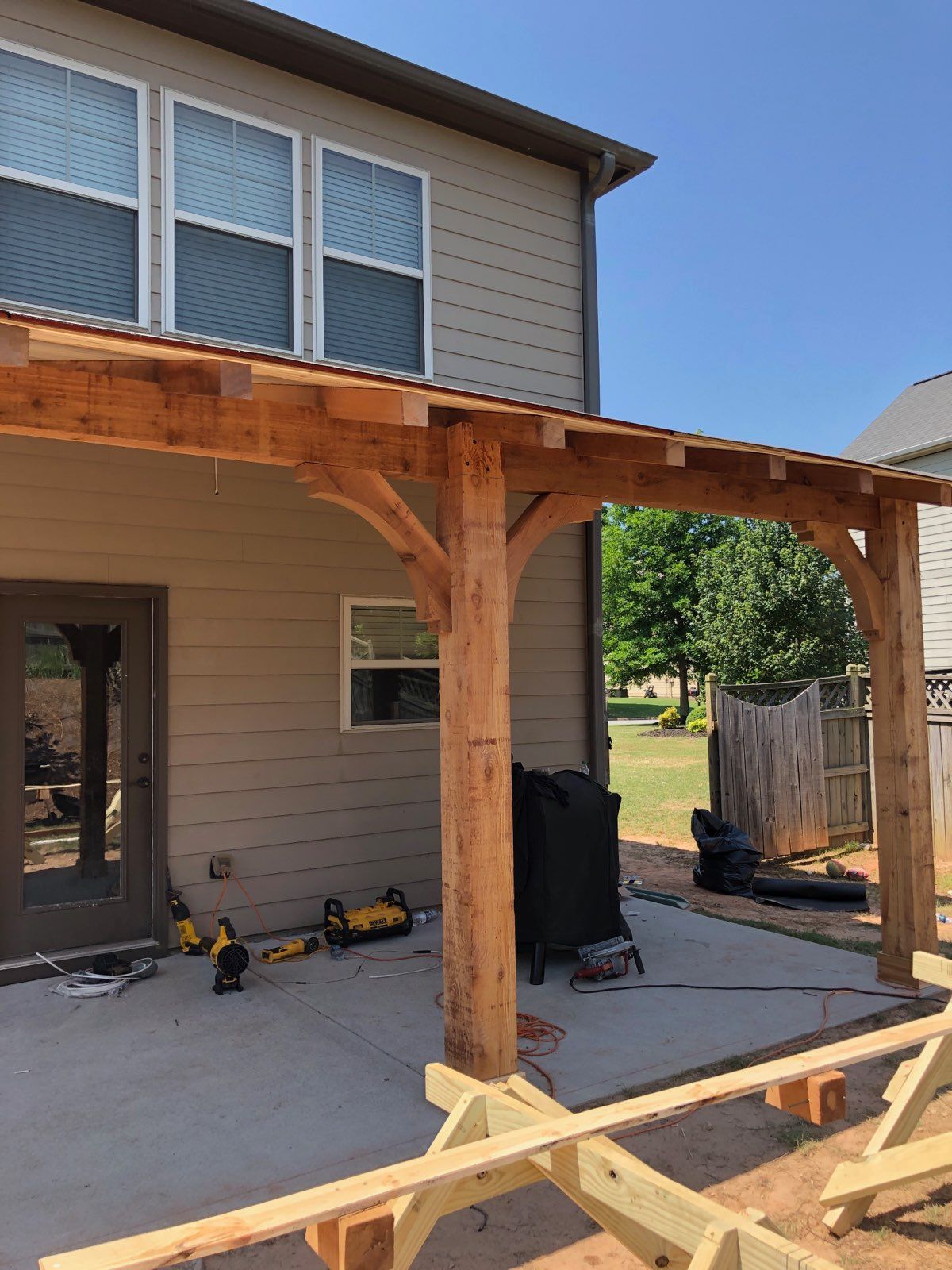 A partially constructed wooden patio cover with posts and roof joists attached to the side of a tan house.