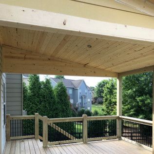 A wooden deck with a covered roof, railings, and black balusters overlooking a backyard with trees and a blue house.