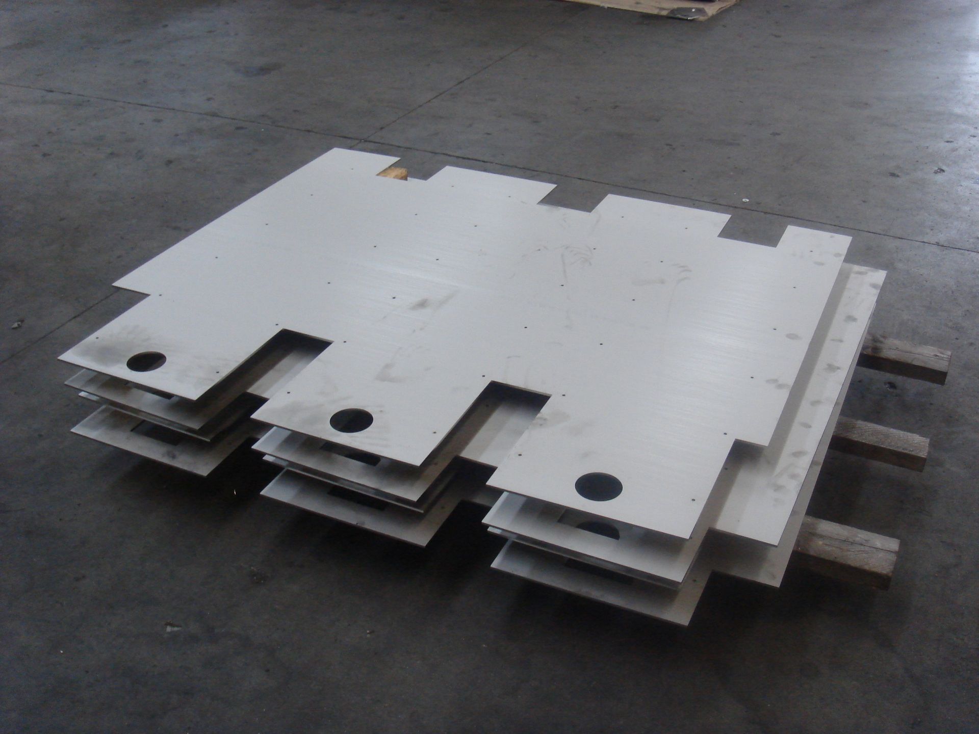 Pannelli di metallo bianco impilati, con linguette e fori ritagliati, appoggiati su un pavimento di cemento su supporti di legno.