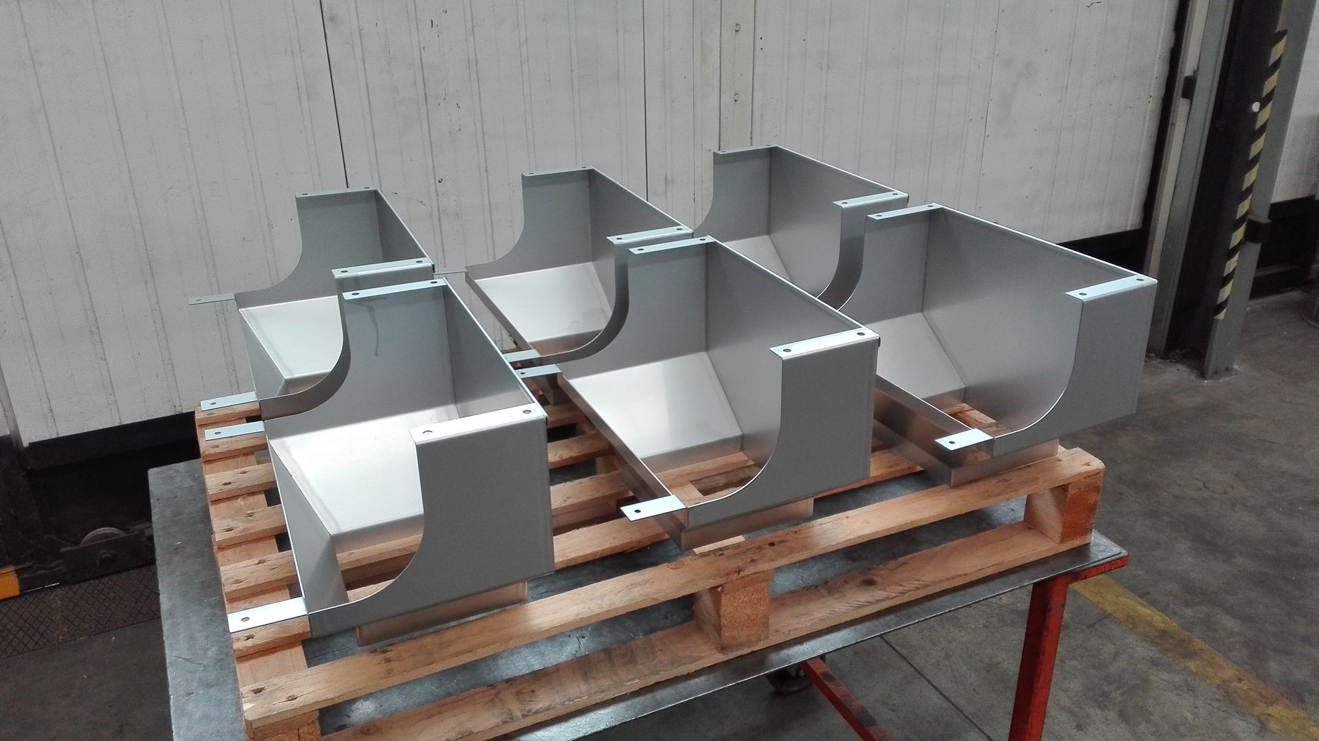 Sei staffe per tubi in metallo argentato impilate su un pallet di legno in un'officina industriale