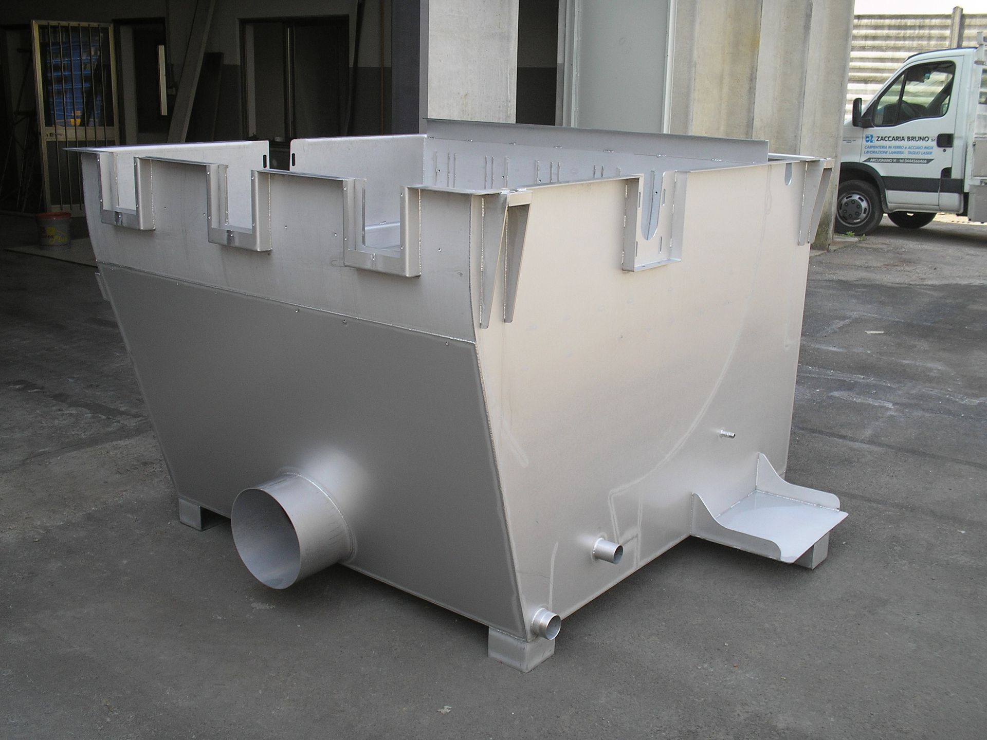 Grande cassonetto industriale in metallo grigio con scarico laterale, mostrato all'aperto vicino a un edificio.