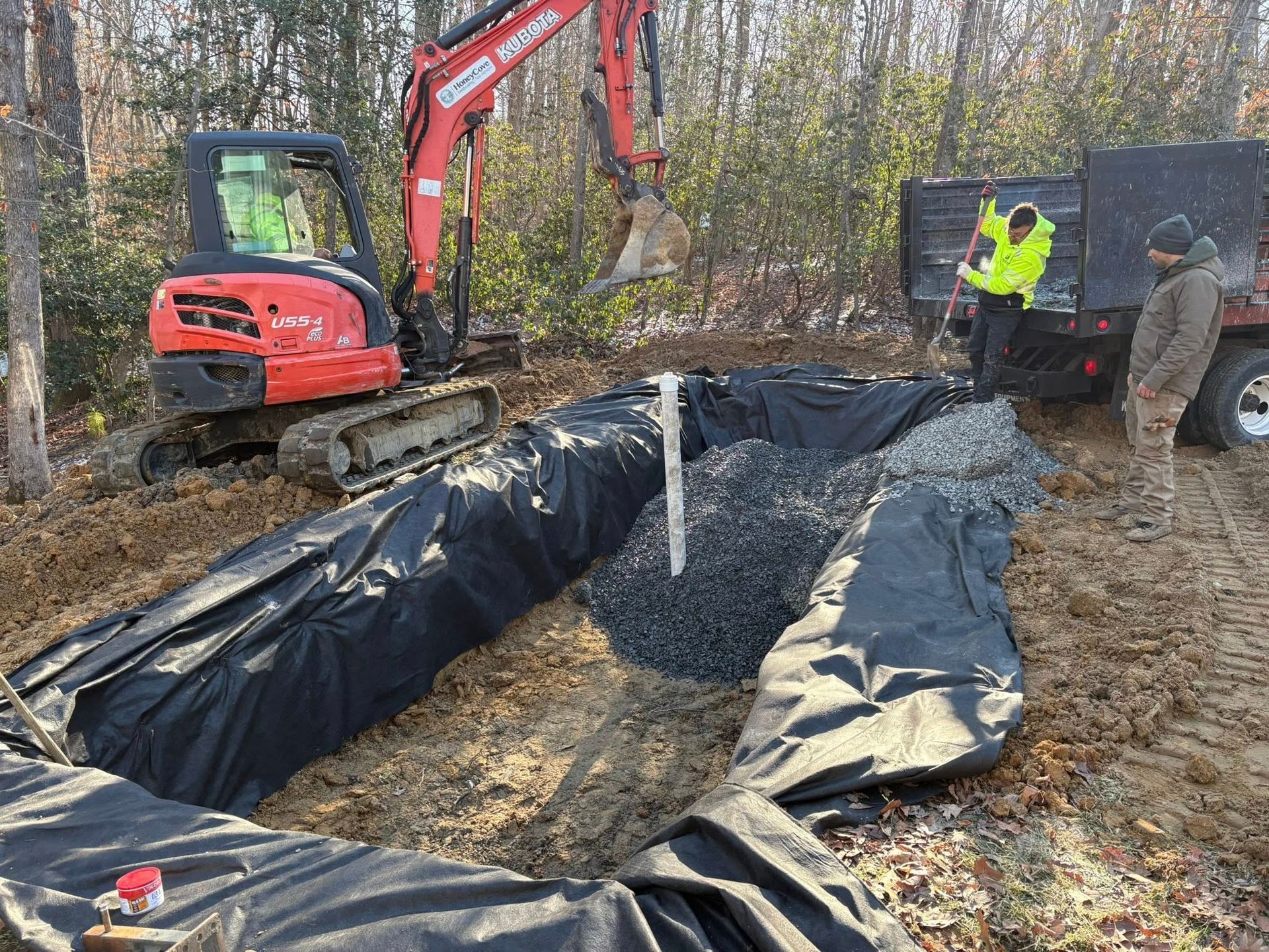 Excavator beside a black-lined pit in a wooded worksite, with workers nearby in safety vests.