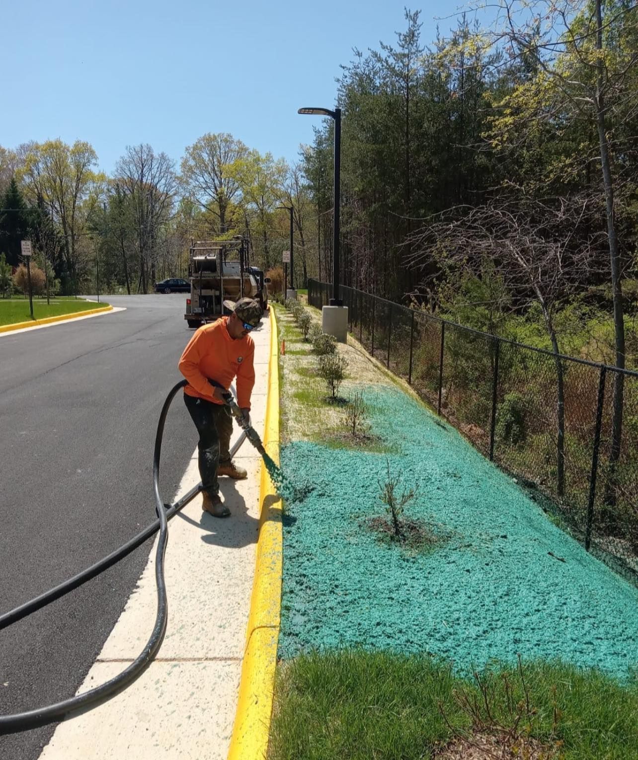 Worker spraying turquoise mulch along a curb beside a road and fenced area