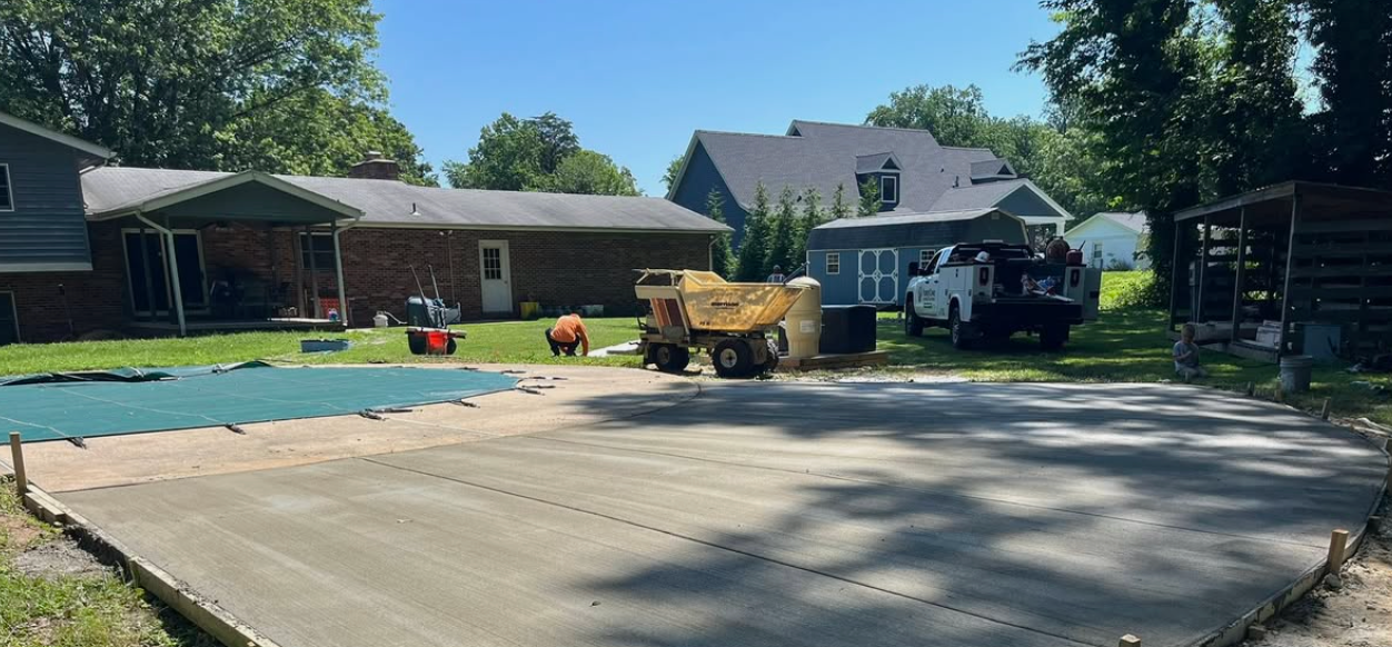 Backyard with an in-ground pool and concrete patio under construction, with a yellow excavator nearby