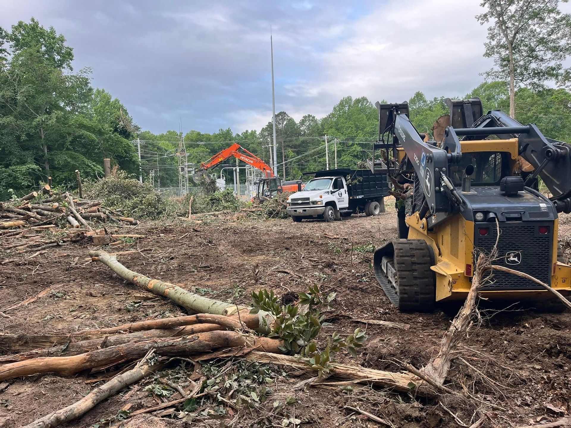 Storm cleanup site with fallen trees, a skid steer, and a white pickup truck under cloudy skies