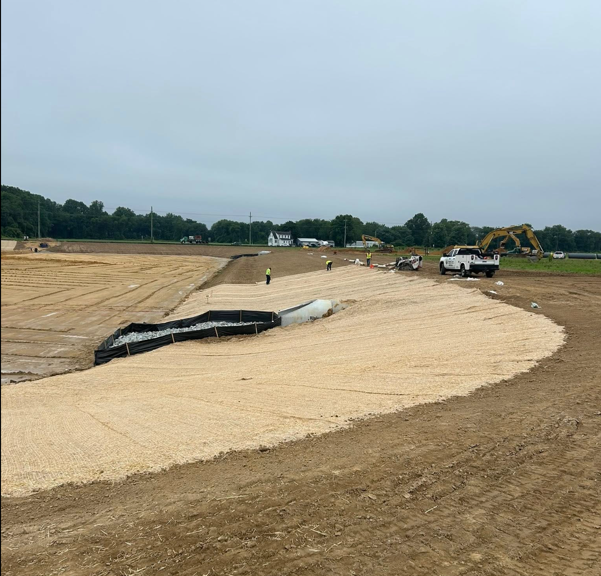 Construction site with gravel road base, excavator, and trucks under an overcast sky
