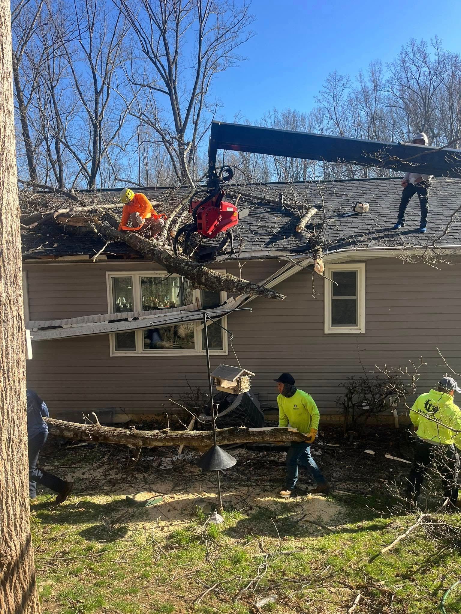 Workers repairing storm-damaged roof on a house, with debris and broken branches around it.