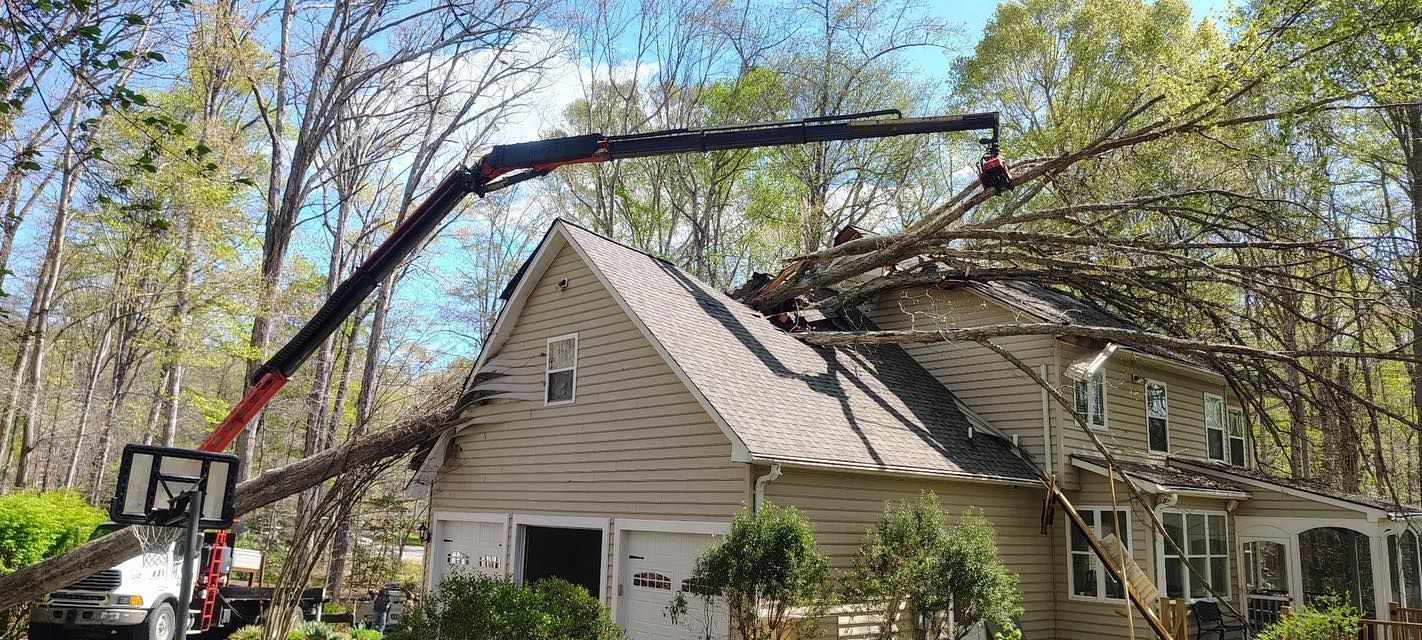 A truck-mounted crane trims tree branches to remove a fallen tree over a beige house.