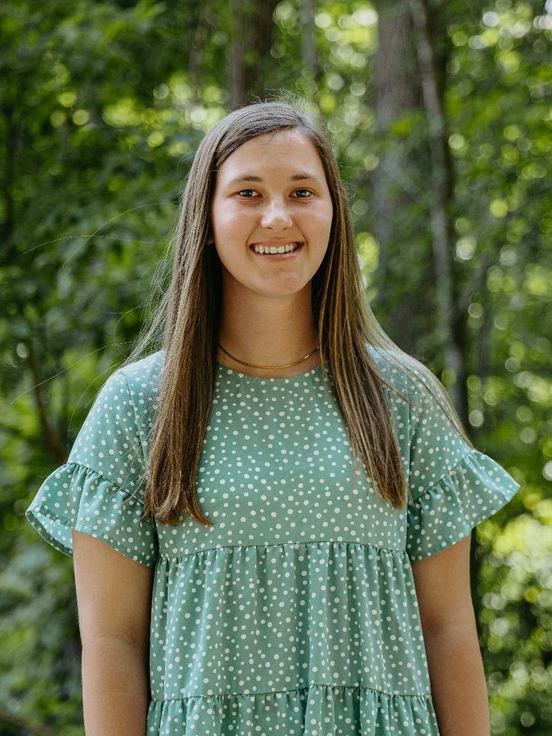 Kaylee in a green dress is standing in front of trees.