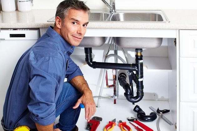 Plumber kneeling under a sink, inspecting pipes with tools on the floor in a kitchen cabinet.