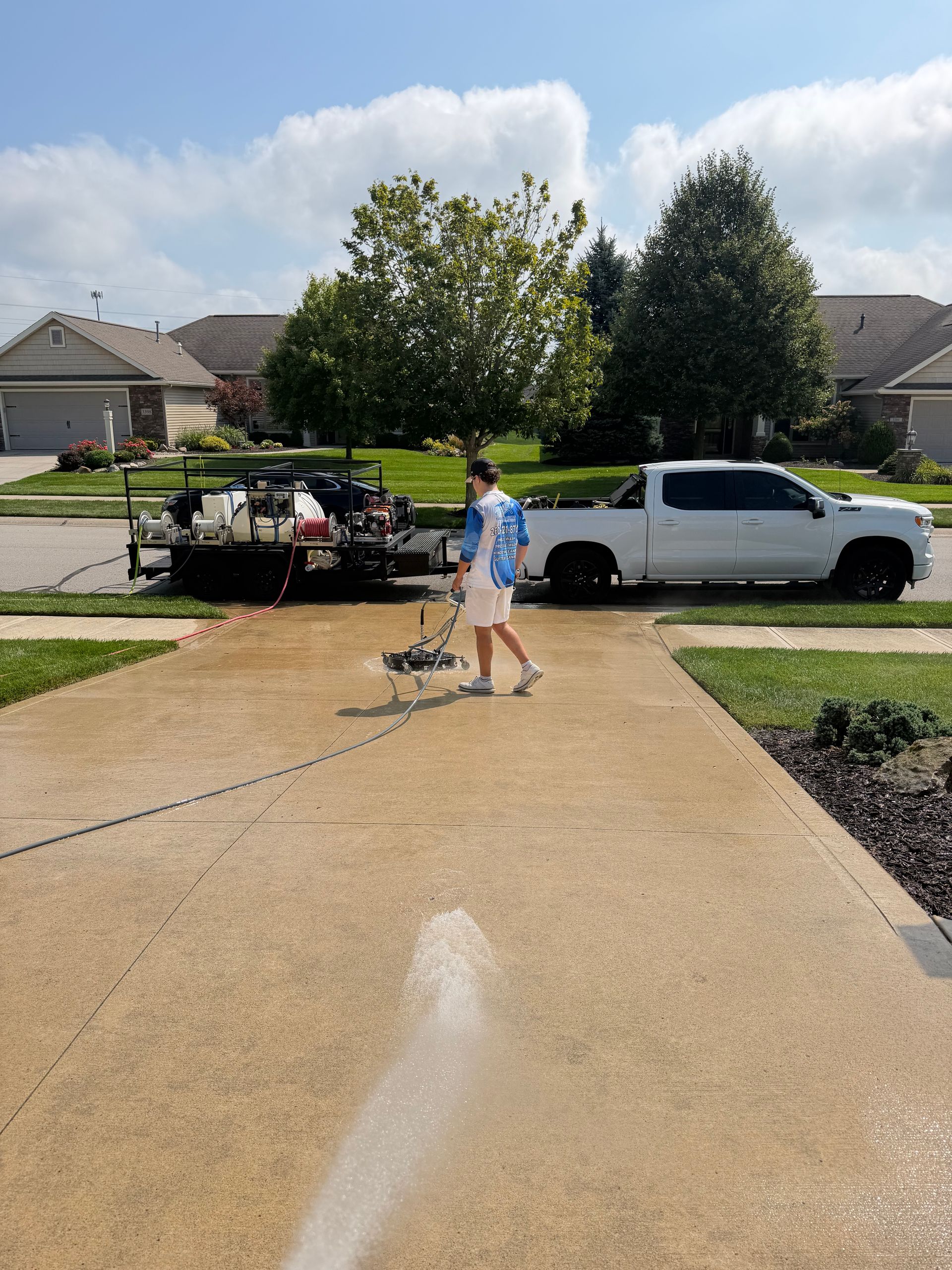 Person washing a driveway with a pressure washer beside a white truck and trailer in a suburban yard