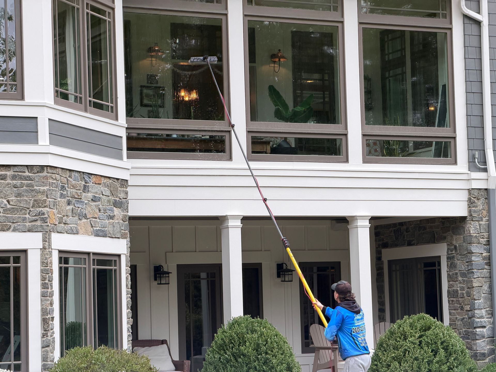 Person in blue jacket power-washing the exterior of a house with large windows and stone siding.