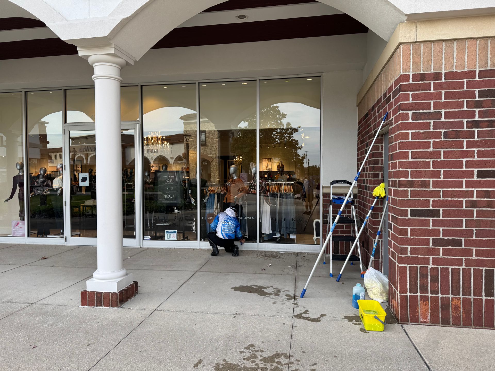 Person seated outside a storefront beside ladders and cleaning supplies at a brick entrance