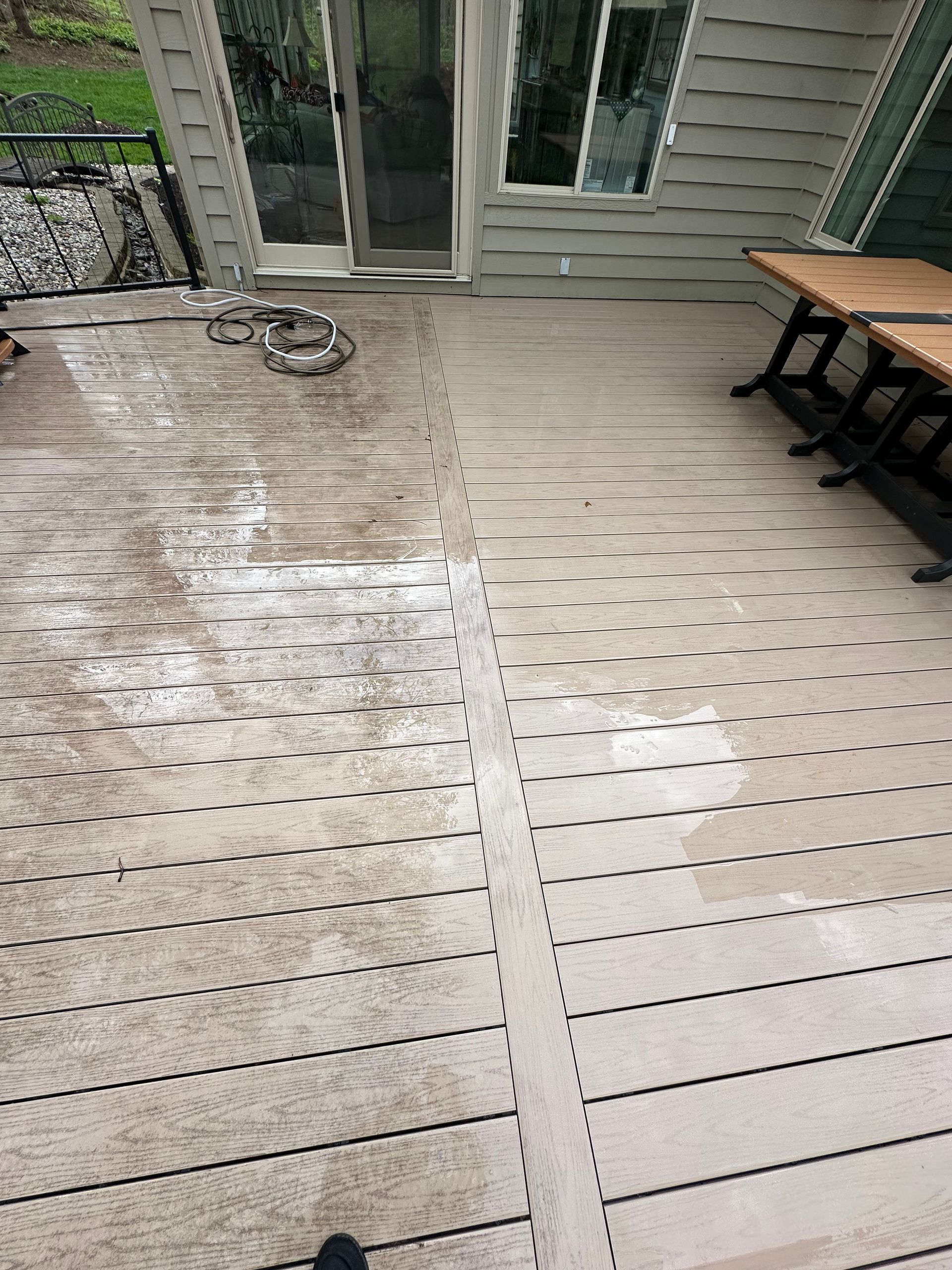 Wooden deck with two damp sections, a coiled hose near glass doors, and a table and bench on the right.